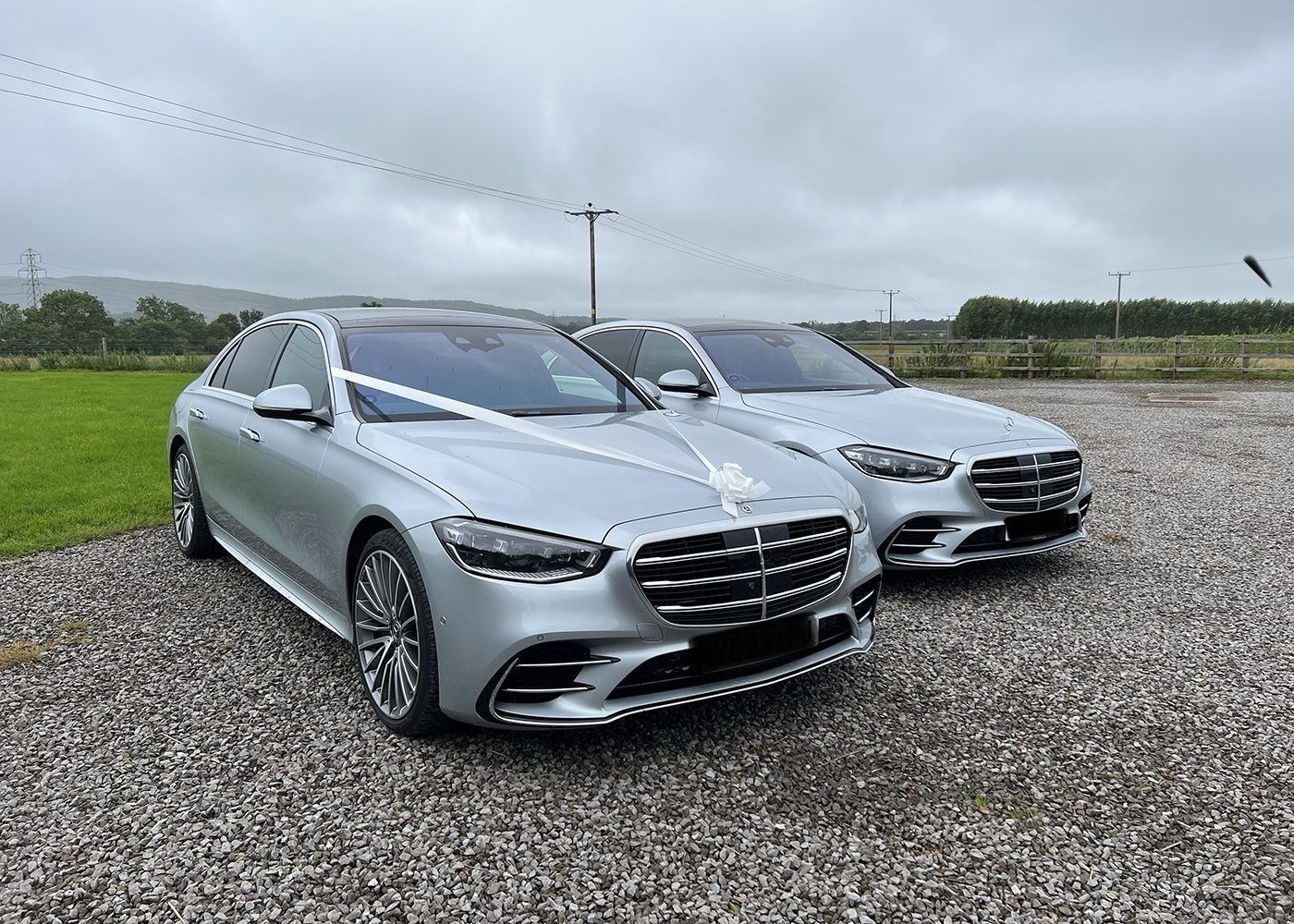 Two silver cars decorated with wedding ribbons on a gravel path with a grassy field in the background.