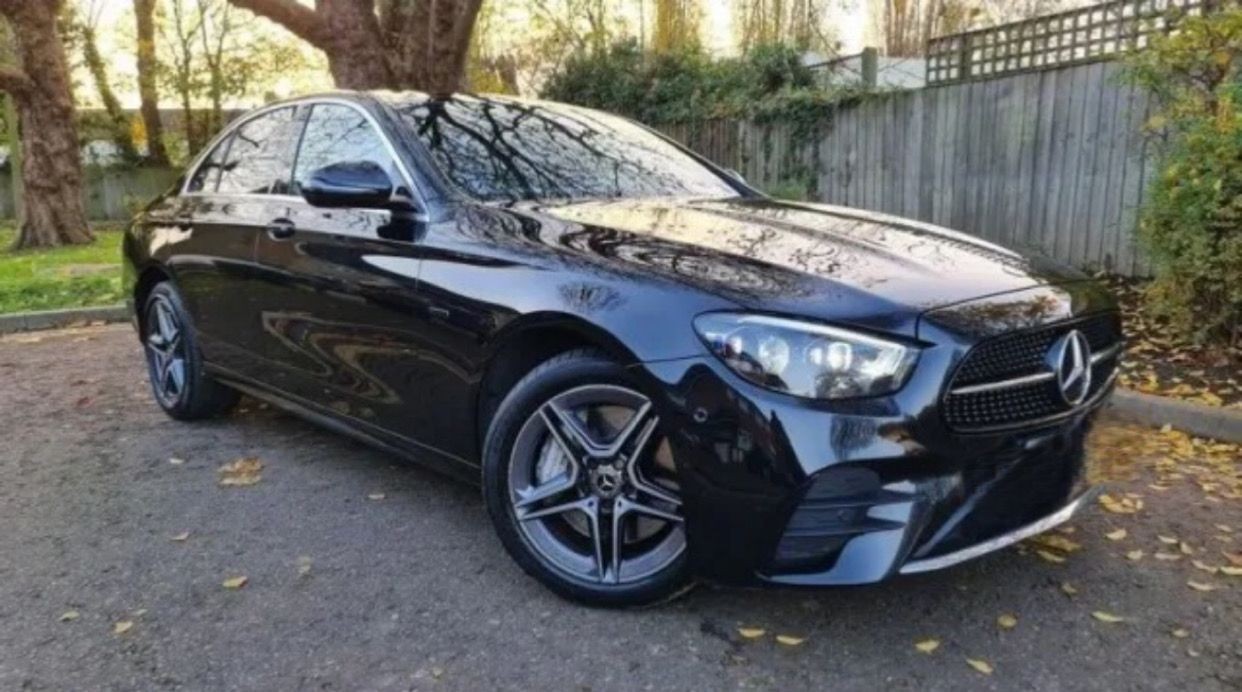 Black luxury sedan parked outdoors on a sunny day with trees and a wooden fence in the background.